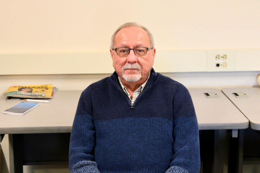 Ricardo, an older man with a small white beard sits in the classroom where he serves as a tutor, wearing a dark blue sweater.